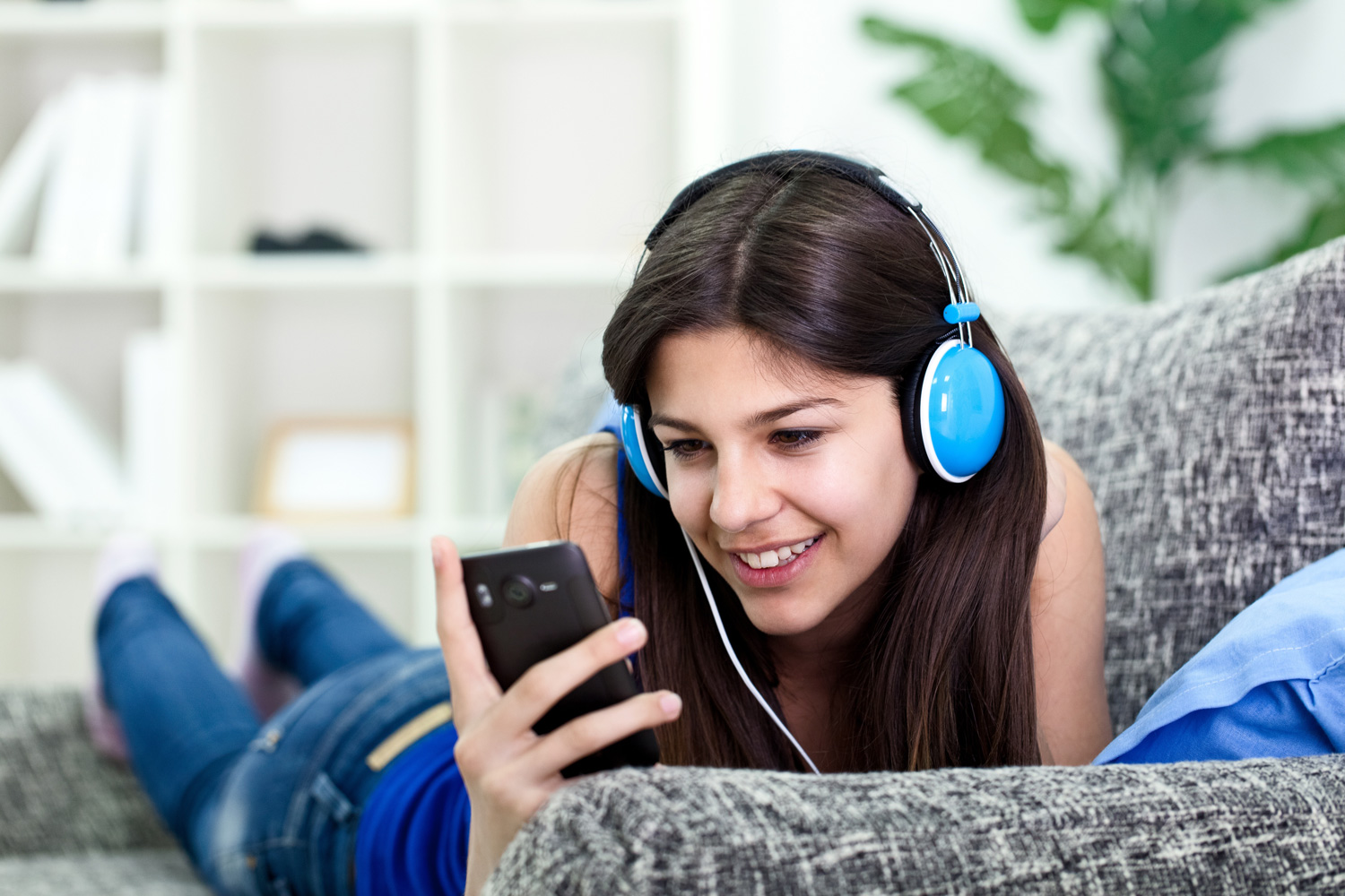 A woman wearing headphones looks at a smartphone while listening to music on a sofa.