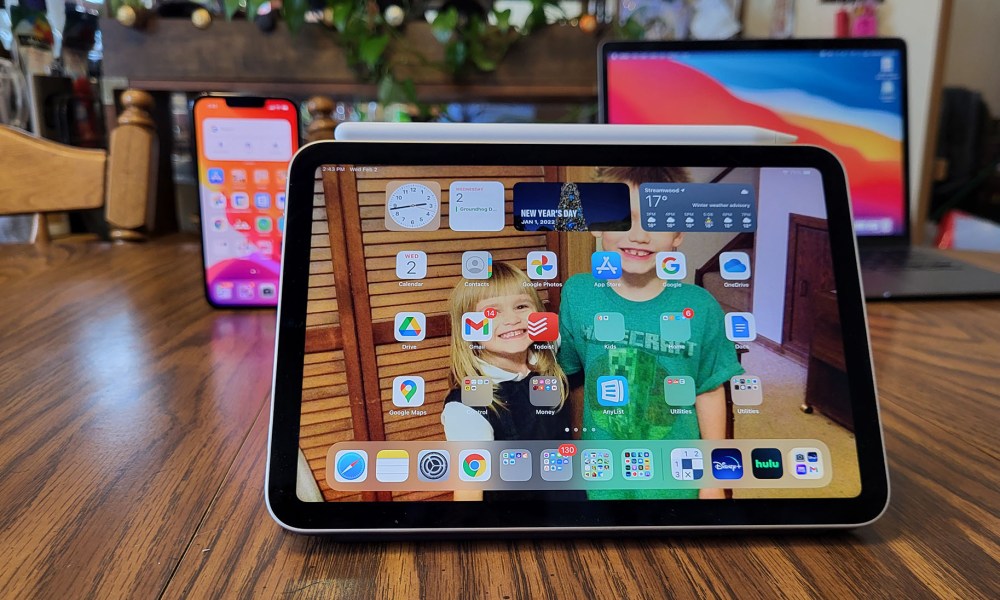 The iPad Mini stands on a floor with other Apple devices in the background.