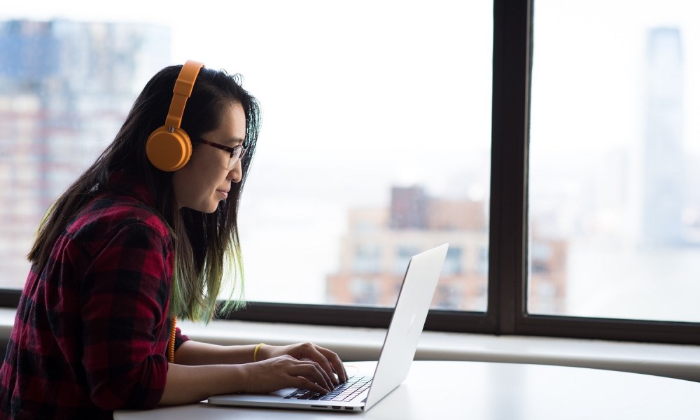 Side view of a person using a laptop while sitting at a table and wearing headphones.