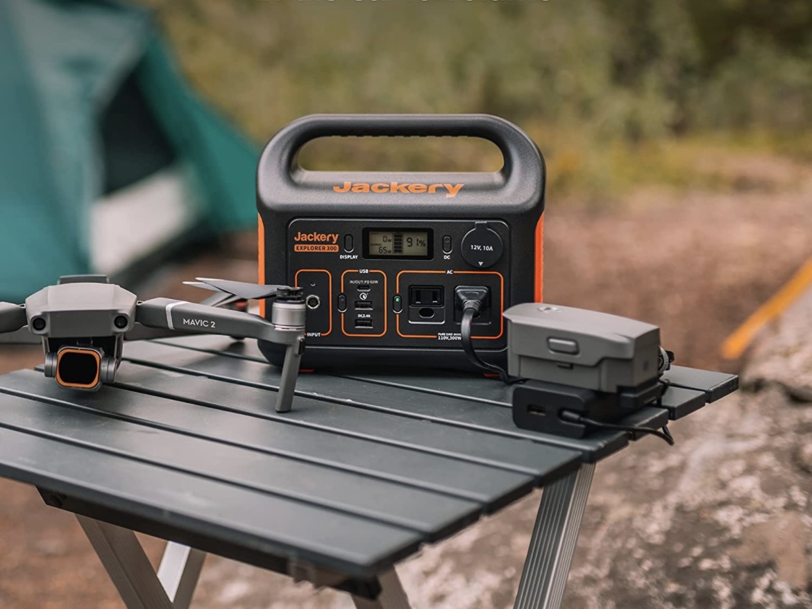 Jackery Portable Power Station Explorer 300 sits on a table at a camping site next to a drone.