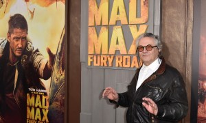 George Miller stands in front of a Mad Max poster.