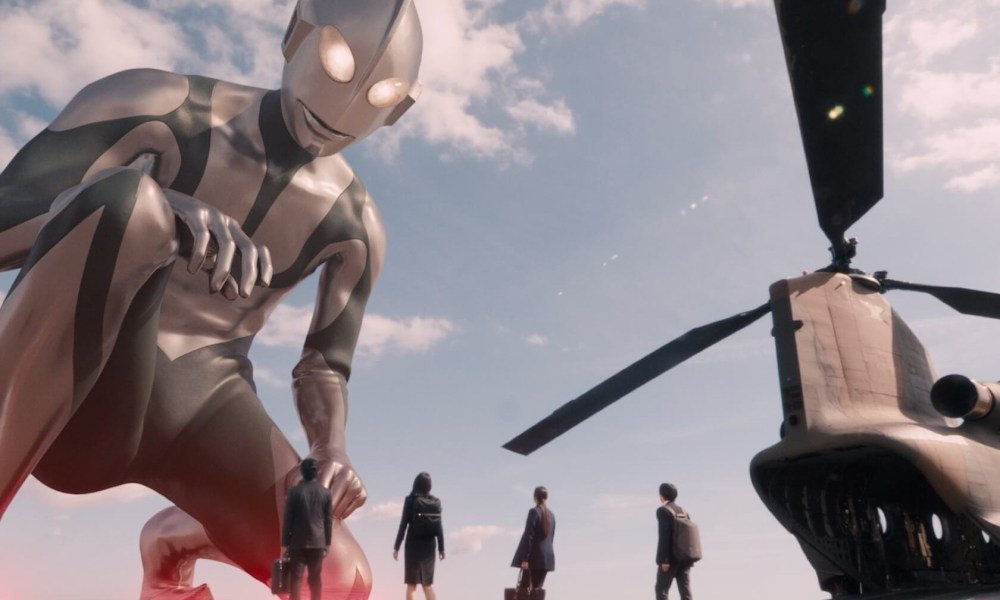 A giant being, Ultraman, bends down to look at four humans on the ground next to a helicopter.