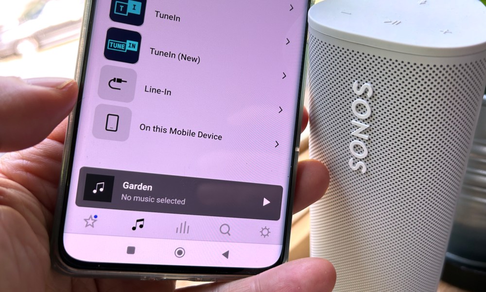 An Android phone with the Sonos app showing the music sources tab menu, next to a Sonos Roam wireless speaker.