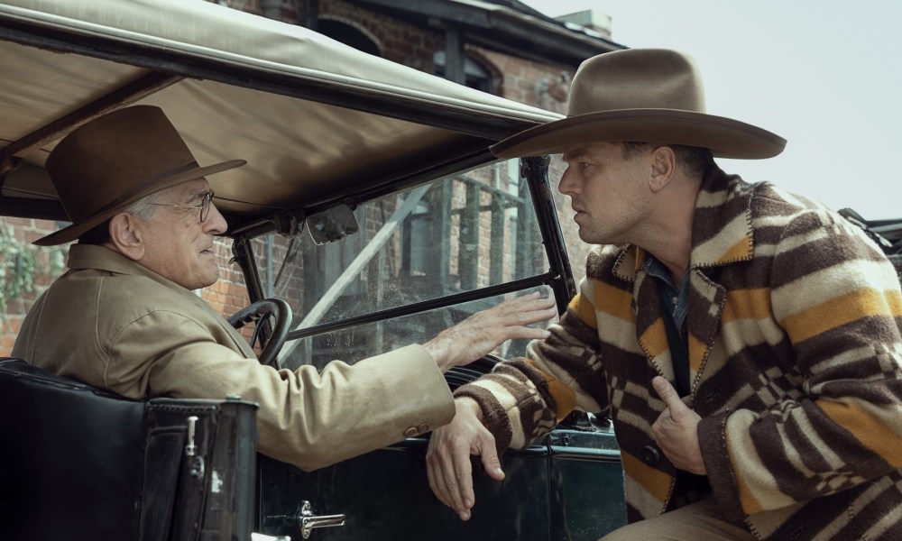 Robert De Niro sits in a car and talks to Leonardo DiCaprio, who leans against it.