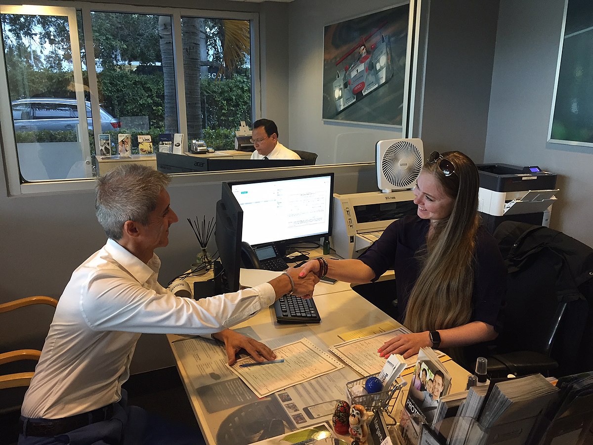 A customer purchases a car at a car dealership and shakes hands with the sales person.