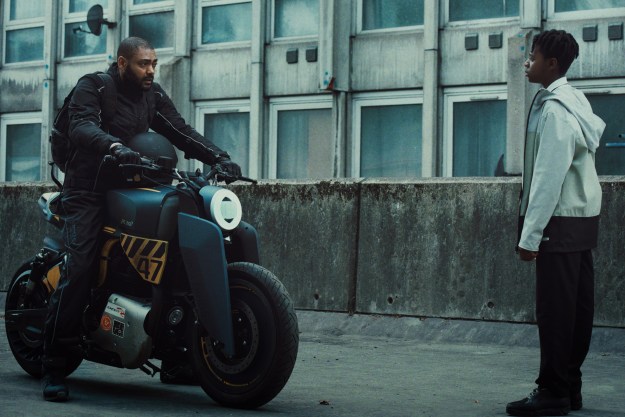 Izi sits on his motorcycle in front of Benji in The Kitchen.