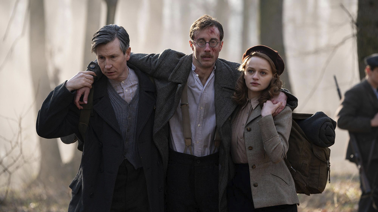 A young man and woman carrying an older, injured man, his arms around their shoulders on either side, in a scene from We Were the Lucky Ones.