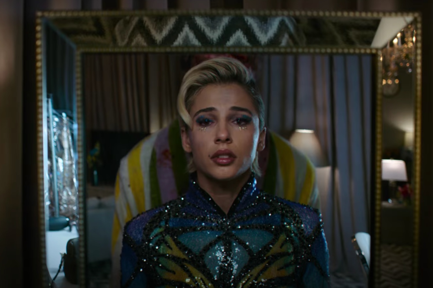 A popstar looks sad while staring into a mirror with someone standing behind her.