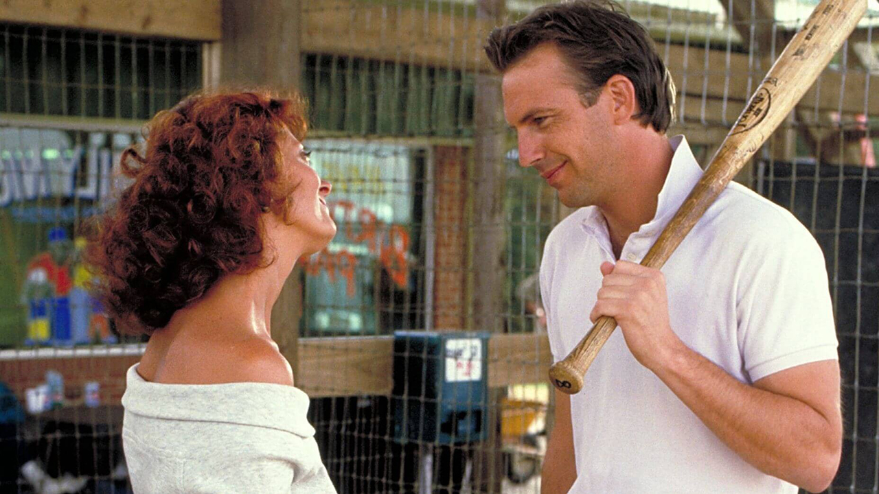 Susan Sarandon looking lovingly at Kevin Costner who is holding a baseball bat in a scene from Bull Durham.