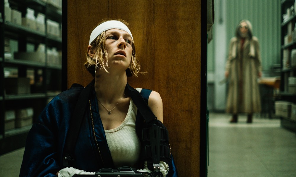 An injured woman sits up against a shelf as another woman stands down the hallway.