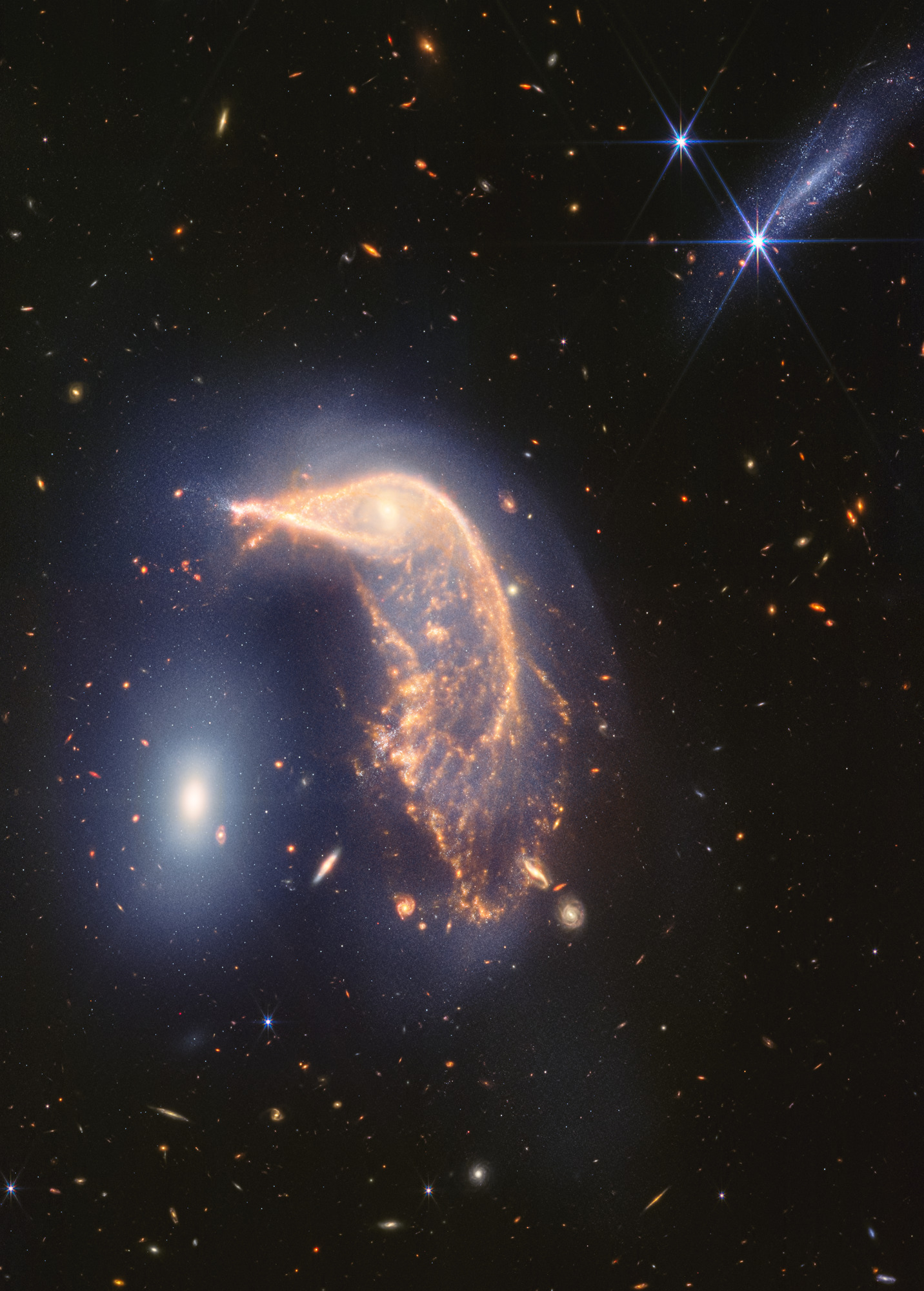 This “penguin party” is loud! The distorted spiral galaxy at center, the Penguin, and the compact elliptical galaxy at left, the Egg, are locked in an active embrace. A new near- and mid-infrared image from the James Webb Space Telescope, taken to mark its second year of science, shows that their interaction is marked by a faint upside-down U-shaped blue glow.