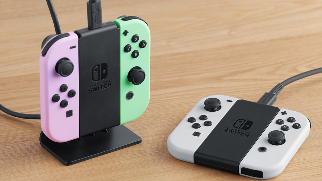 Purple and green Joy-Cons on the black charging stand. There's another charging stand next to it, but it's lying flat on the table with two white Joy-Cons.