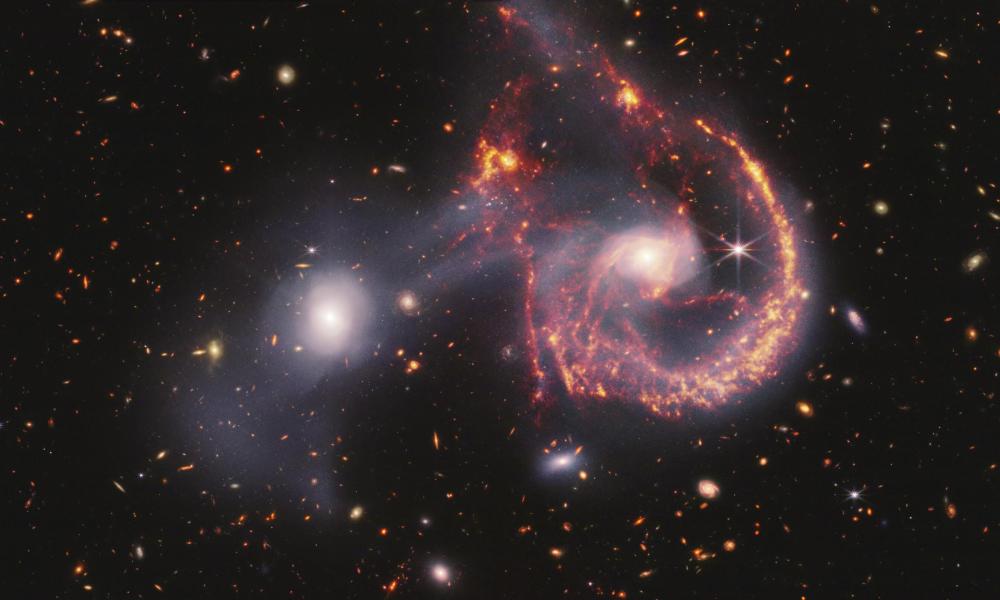 This composite image of Arp 107, created with data from the James Webb Space Telescope’s NIRCam (Near-InfraRed Camera) and MIRI (Mid-InfraRed Instrument), reveals a wealth of information about the star formation taking place in these two galaxies and how they collided hundreds of million years ago. The near-infrared data, shown in white, show older stars, which shine brightly in both galaxies, as well as the tenuous gas bridge that runs between them. The vibrant background galaxies are also brightly illuminated at these wavelengths.