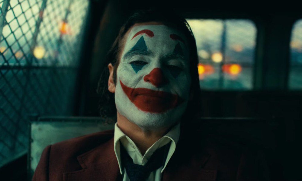 Joaquin Phoenix in full clown makeup sits in the back of a police cruiser in a still from the movie "Joker: Folie à Deux."