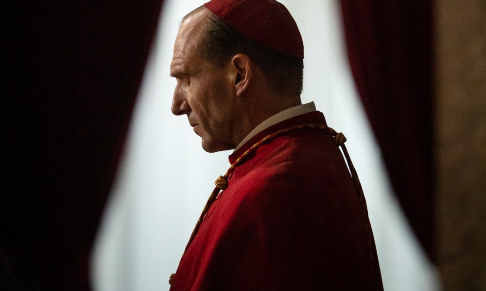 Ralph Fiennes stands next to a window in Conclave.