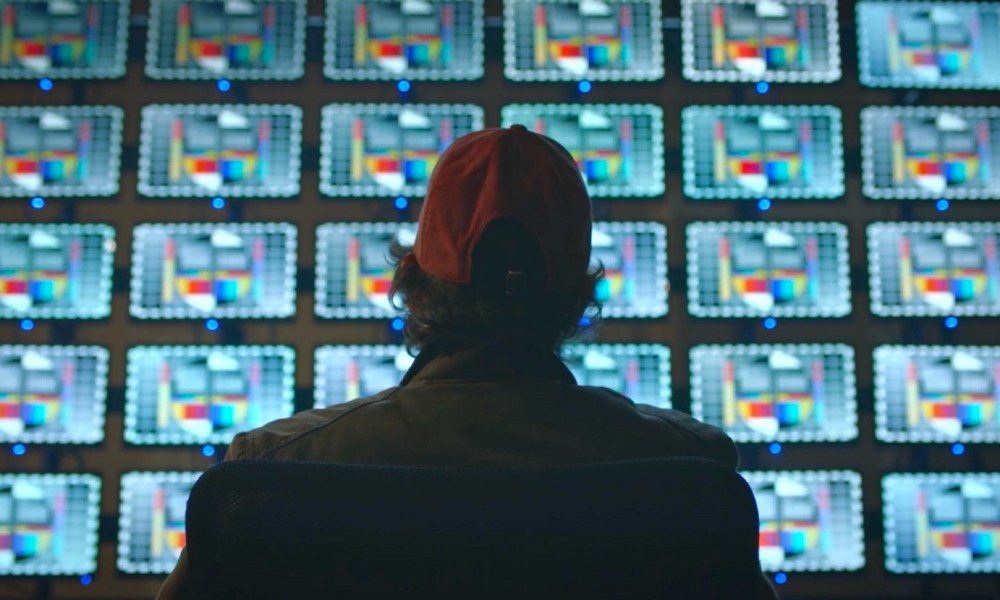 Tim Blake Nelson sits in front of a wall of TV screens in Watchmen.