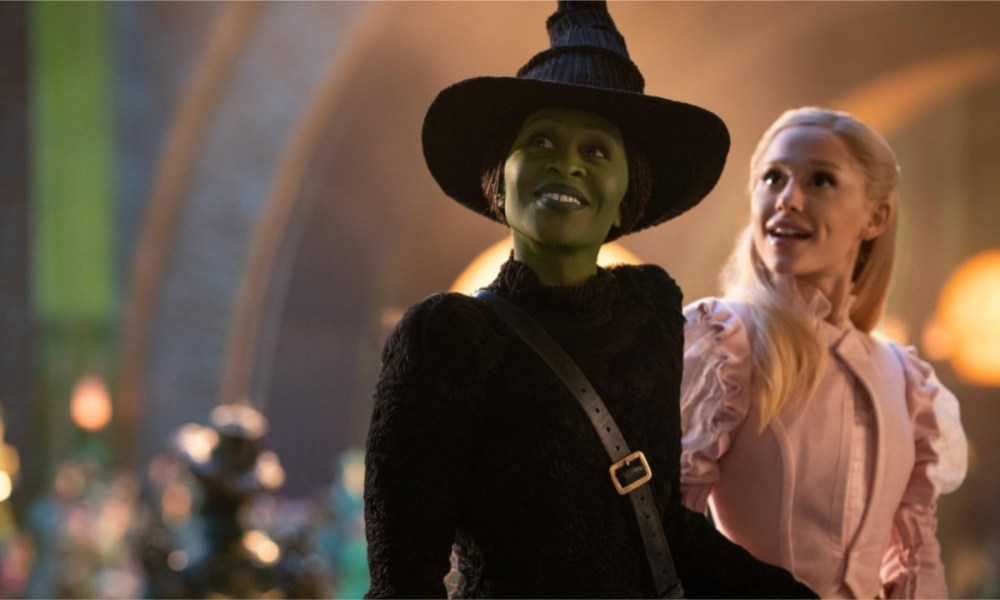 A green witch and a female witch stand next to each other and smile.