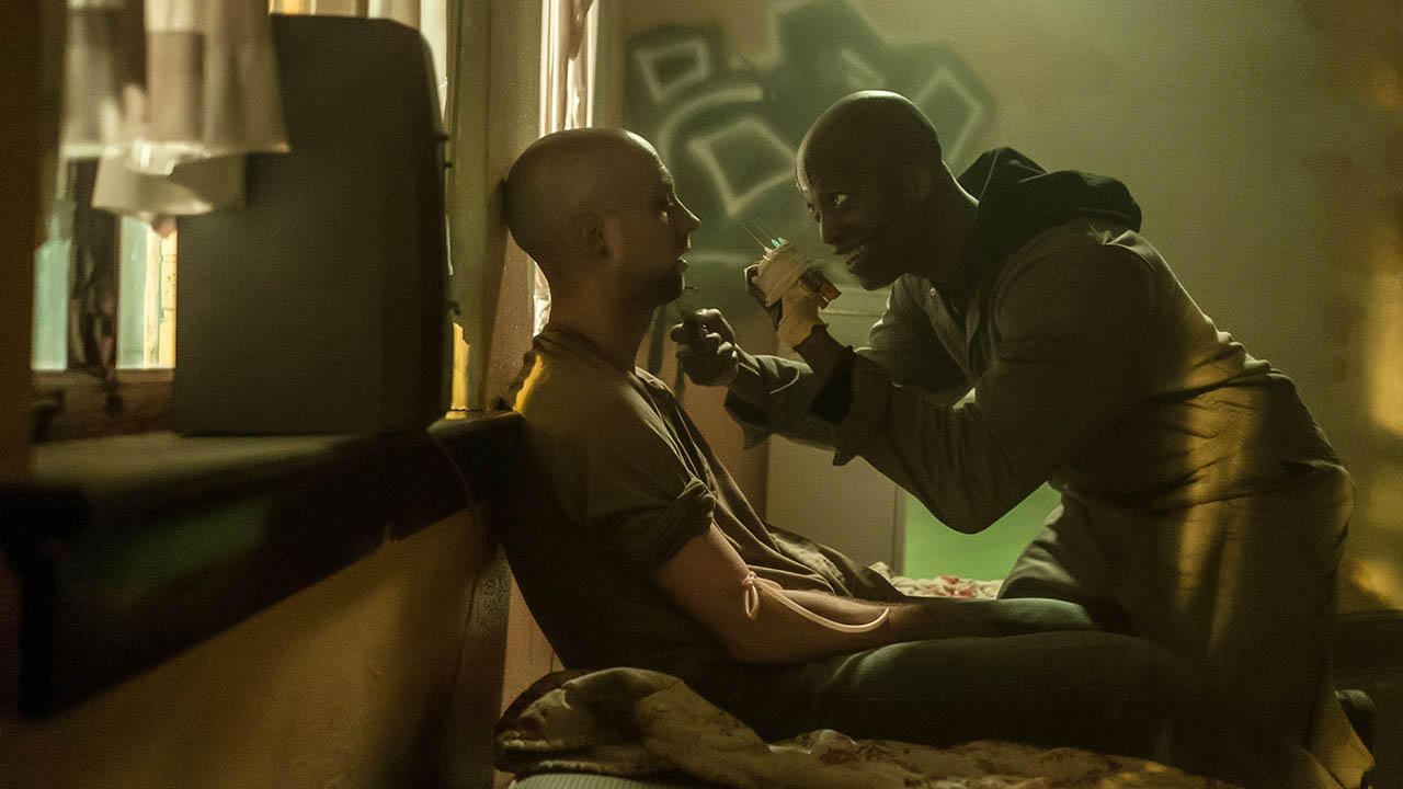 A man holding up a weapon to another man sitting up on a bed in The First Purge.